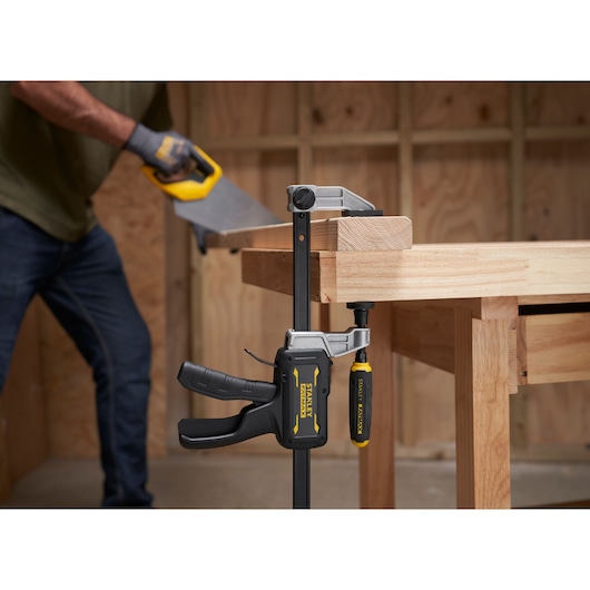 STANLEY® FATMAX® Hybrid Trigger Clamp, 300mm clamping plank of wood to workbench in foreground; wood is sawed in background
