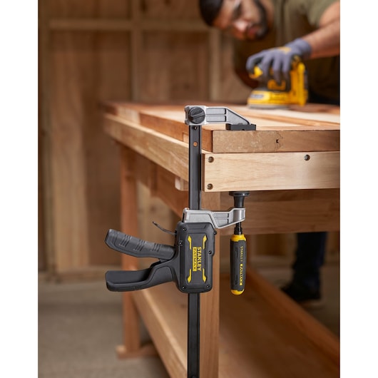 STANLEY® FATMAX® Hybrid Trigger Clamp, 300mm clamping plank of wood to a workbench, front view, while man works in background