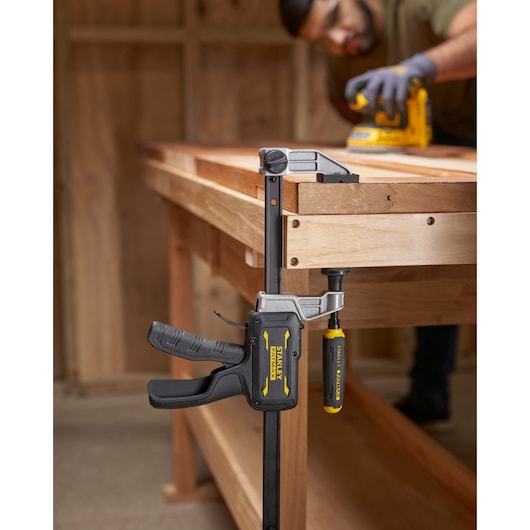 STANLEY® FATMAX® Hybrid Trigger Clamp, 450mm clamping plank of wood to a workbench, front view, while man works in background