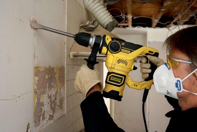 Photo d'une personne utilisant le marteau rotatif STANLEY FME1250K, perçant un mur avec gants et masque de sécurité.