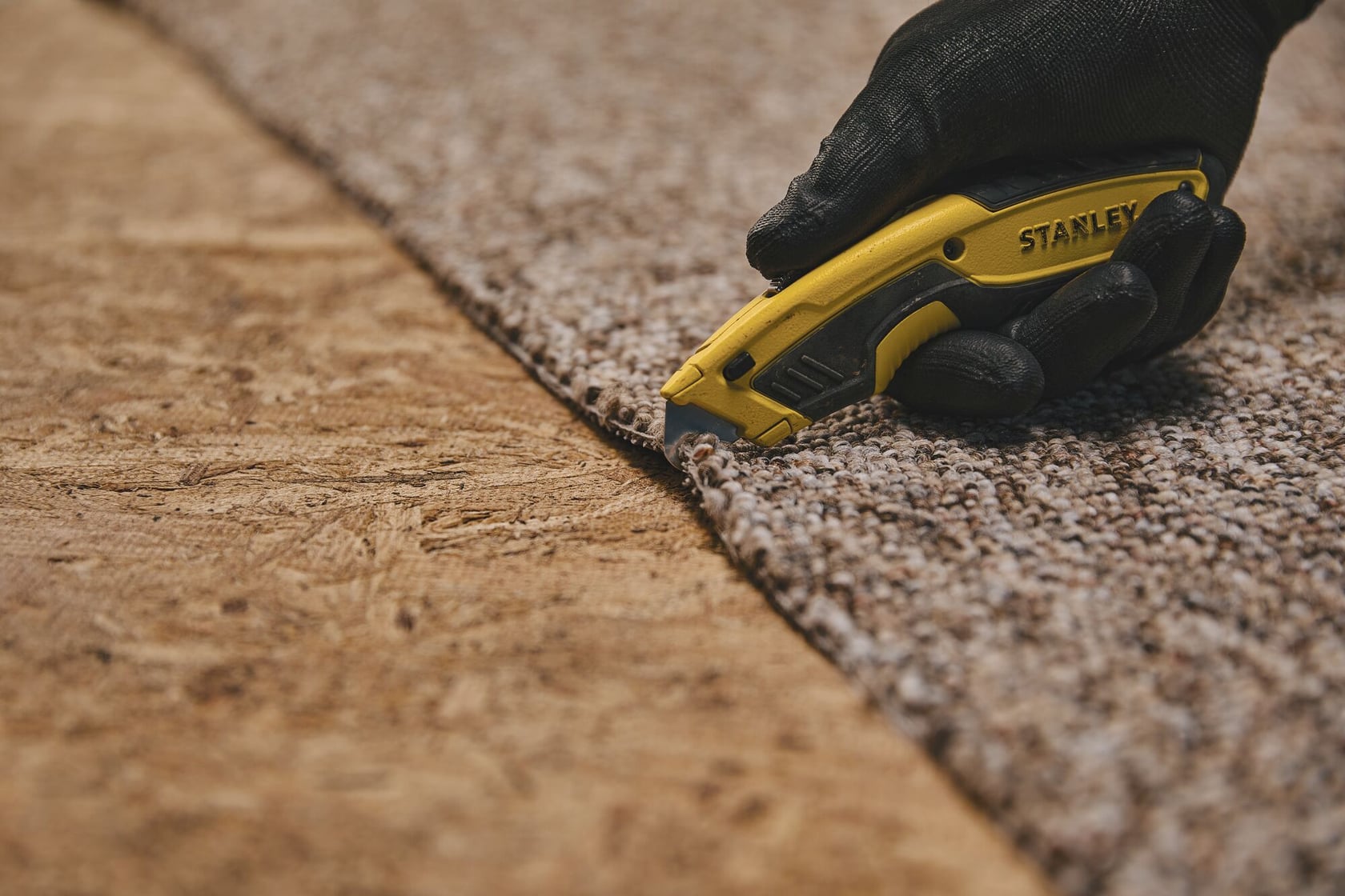 Cutter Stanley STHT10479 découpant de la moquette, gros plan, utilisateur avec gants noirs.