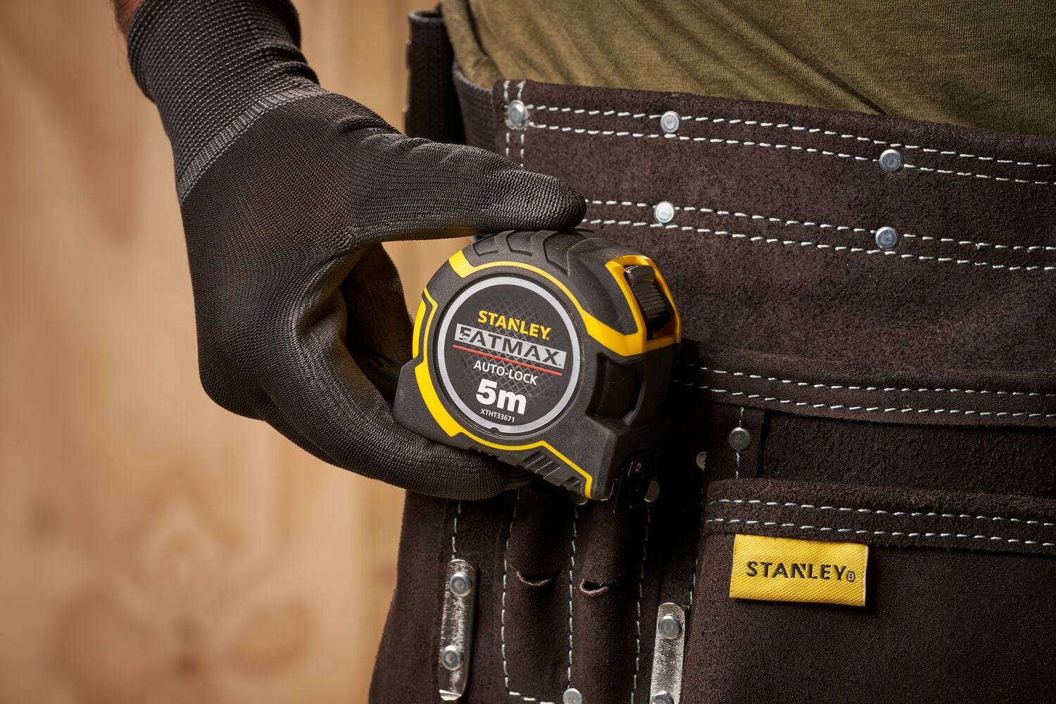 STANLEY XTHT0-33671 mètre ruban tenu par une main gantée, attaché à une ceinture à outils, vue rapprochée.